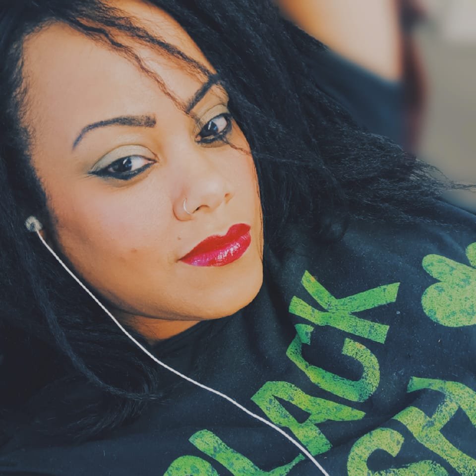 A woman with long dark hair, bold red lipstick, and a nose ring wearing wired earbuds and a black shirt with green text